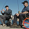 Image of two people sitting on a seaside-jetty with fishing rods. One has a bag of supermarket prawns next to them; there is a large red circle with a line through it superimposed on the bag of prawns.