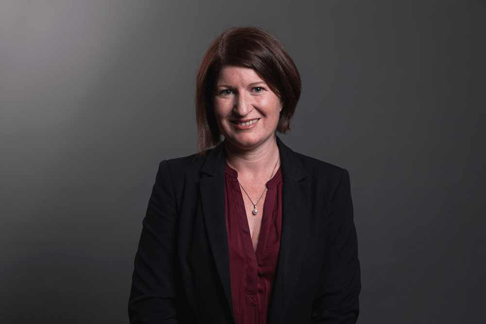 Image of a smiling person with short brown hair, wearing a read top and black blazer. They are standing in front of a grey backdrop.