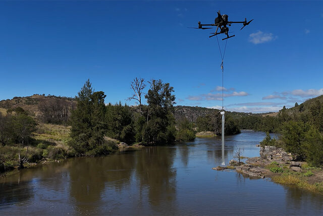 Image of a drone flying over a river surrounded by greenery, collecting a water sample