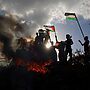A group of people who demonstrates with flags, smoke and flames around them and blue sky with some clouds and sun.