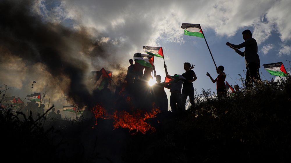 A group of people who demonstrates with flags, smoke and flames around them and blue sky with some clouds and sun.