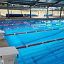 Sparkling partially-undercover in-ground public swimming pool and pool deck, with pool covers on rolls in background and flags above the water surface
