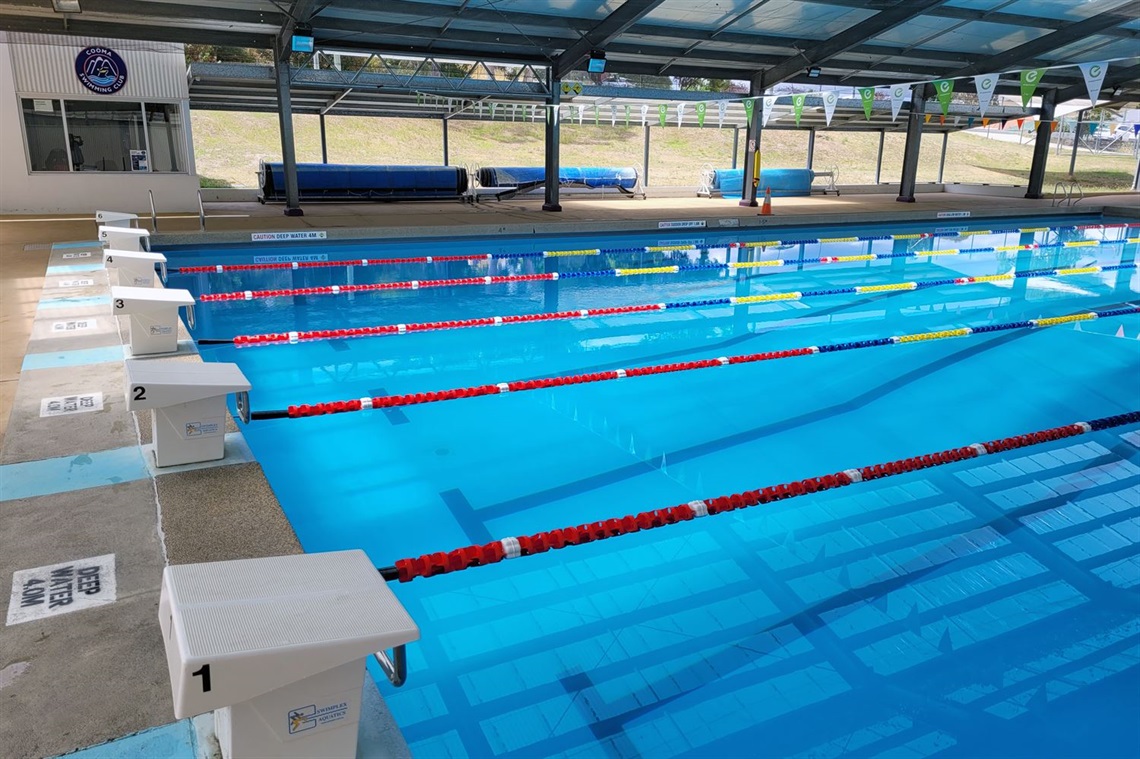 Sparkling partially-undercover in-ground public swimming pool and pool deck, with pool covers on rolls in background and flags above the water surface