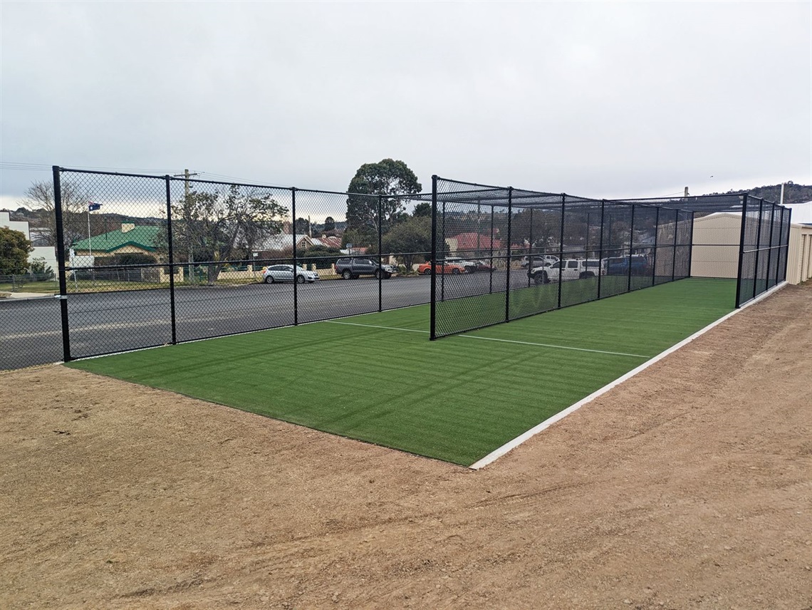 New black wire-mesh fencing around fresh artificial turf surrounded by freshly seeded earth with Bombala street scene in background