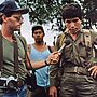 A reporter with a camera hanging from his neck and microphone in his hand interviews a military officer.