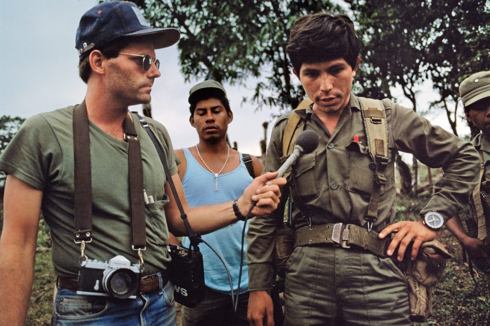 A reporter with a camera hanging from his neck and microphone in his hand interviews a military officer.