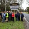 Picture of a group of men and women in front of a new sign that warns that the road ahead is flooded. The picture is taken on the side of the road and the weather is wet.