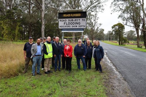 Picture of a group of men and women in front of a new sign that warns that the road ahead is flooded. The picture is taken on the side of the road and the weather is wet.