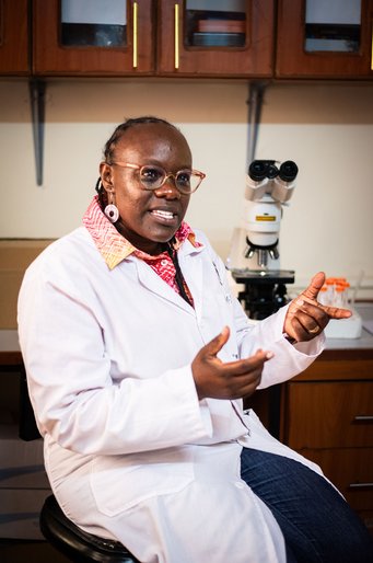 A person wearing a white lab coat gestures while seated, with a microscope and laboratory equipment visible in the background