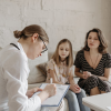 A healthcare professional sits with a clipboard, taking notes while speaking with a woman and a child seated together on a sofa in a softly lit room, suggesting a supportive consultation setting