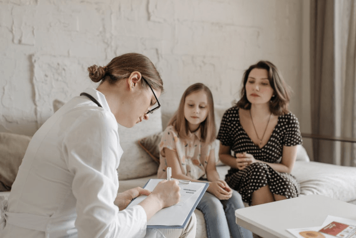 A healthcare professional sits with a clipboard, taking notes while speaking with a woman and a child seated together on a sofa in a softly lit room, suggesting a supportive consultation setting